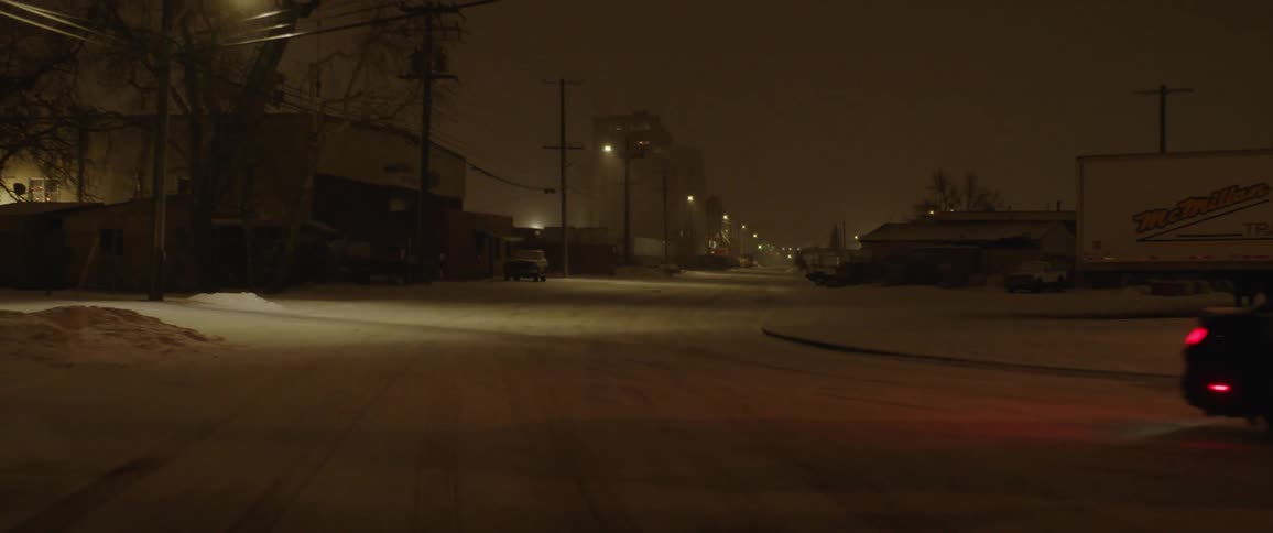 Car drives down street at night in snow under power lines and past buildings trees and street lights