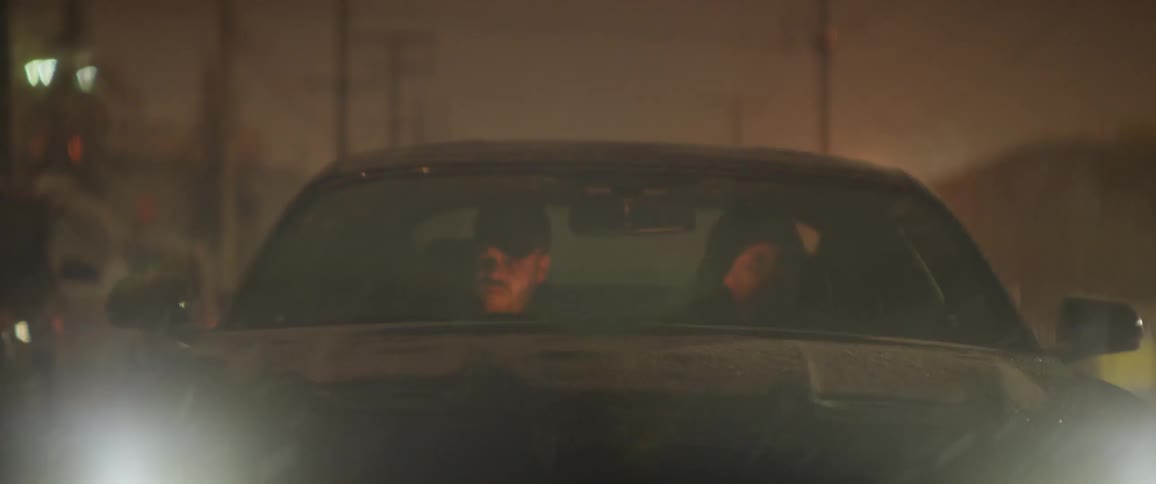 Two people in a car driving down a street as snow falls at night