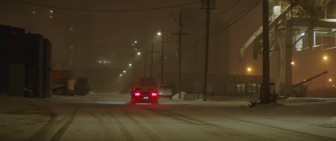 Cardrives down snow covered road at night past power lines and a metal tower and brakes as it comes close to a few trucks