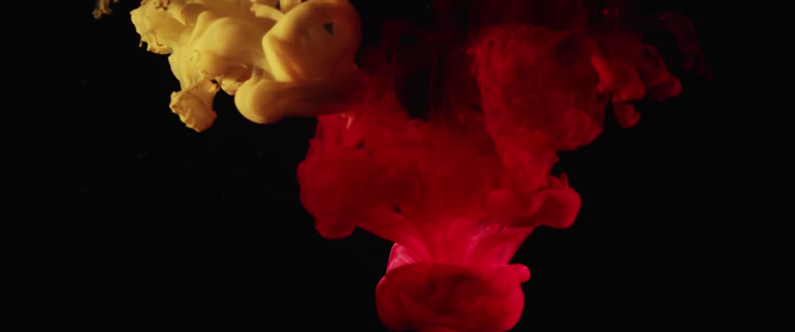 A splash of red and yellow paint fall into what appears to be water and create smoky patterns