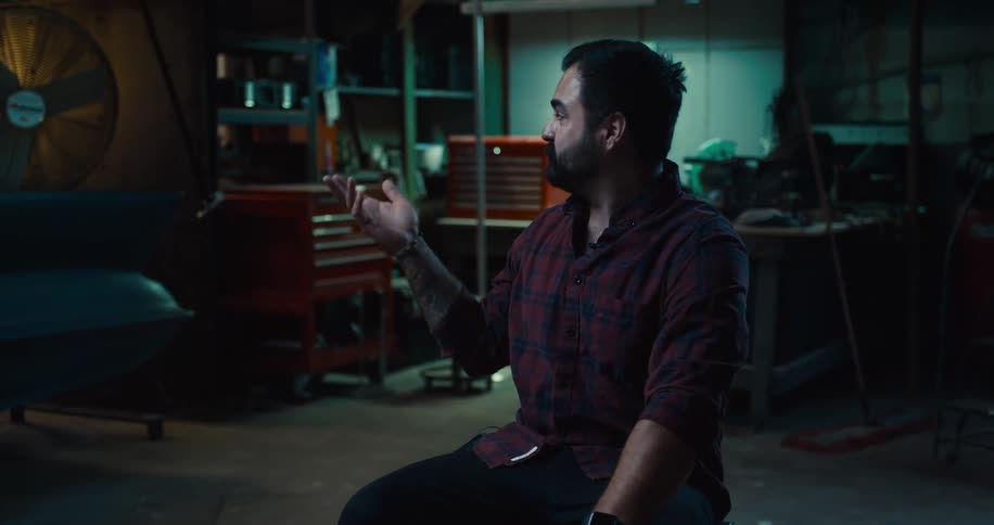 A man sits on a stool talking and gesturing while looking at his dark work space with red tool cabinets and a large fan