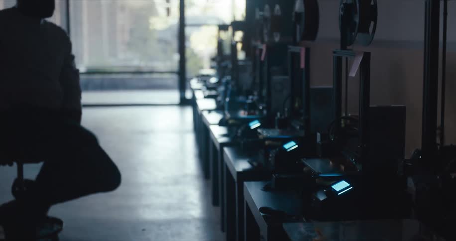 Man sits on a stool with wheels and pushes himself across the floor in front of a row of electronics