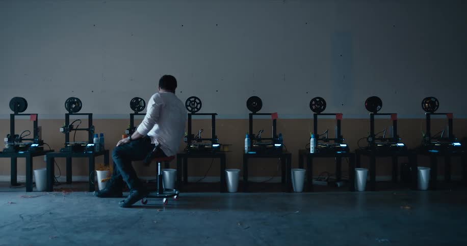 Man slides past row of similar machines on stool stopping to inspect one