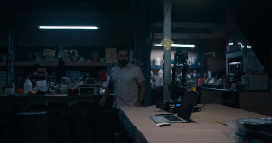 A man stands next to a car and then walks in to a workshop and looks at a laptop