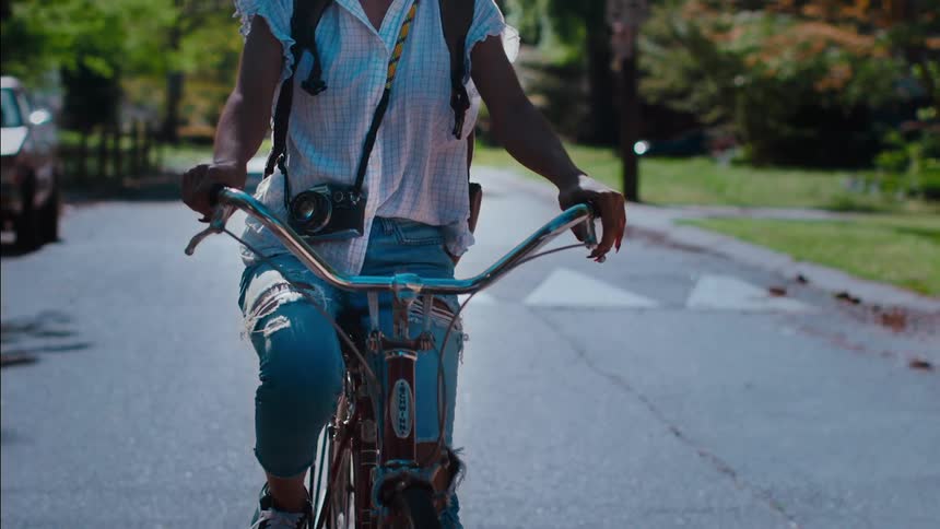 A female photographer rides a bicycle along a road through a neighborhood on a sunny day