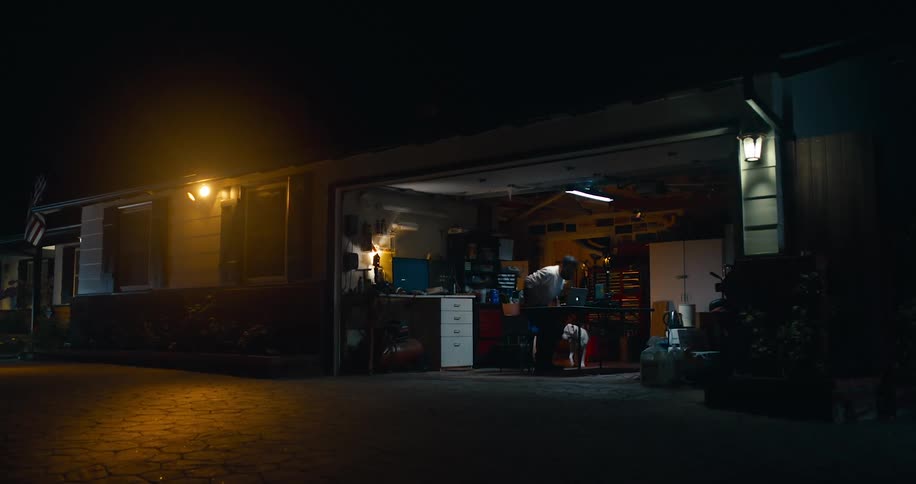A man sits down at his computer in his garage and pets his dog