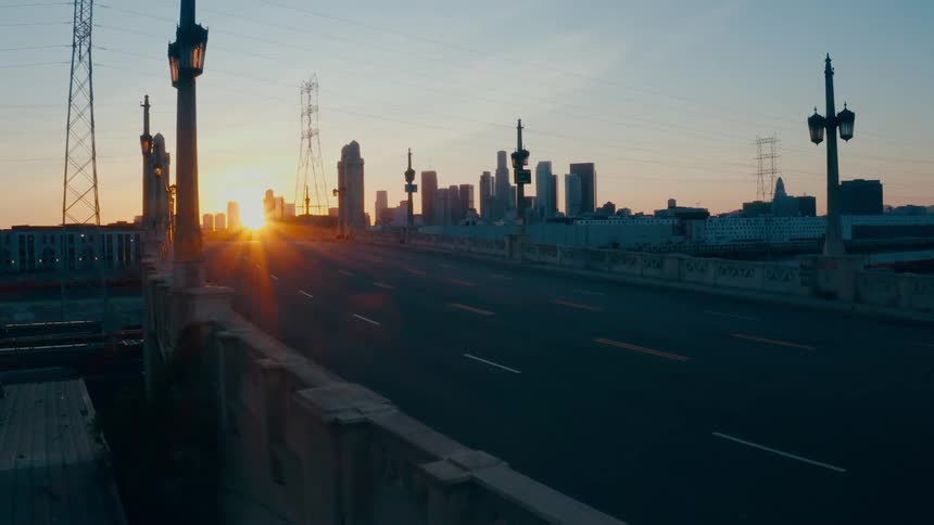 A sun sets over a city skyline and a bridge with a road leading into the city contains a set of street lamps