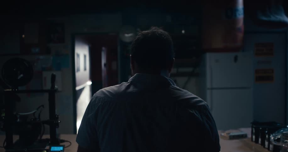 A man at a table with a 3d printer with a doorway and a refrigerator in the background