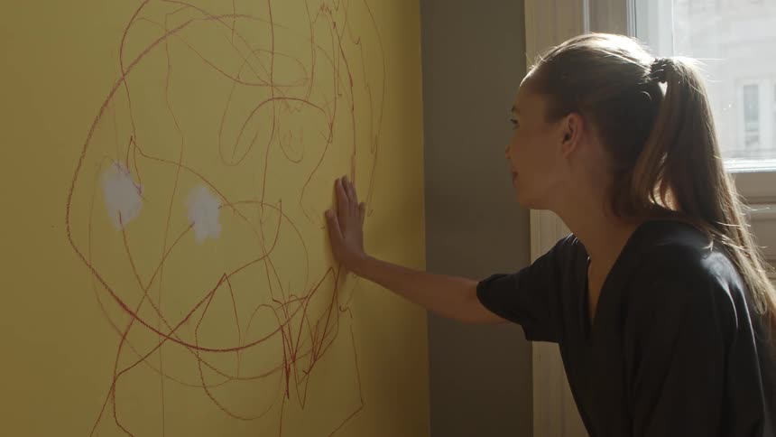 A woman bends down and looks at the scribbling on a wall in her home