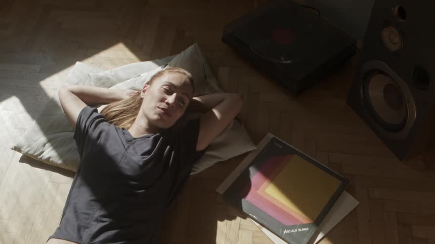 Woman lays in the floor on pillows in a square of sunlight beside some books and papers with speakers to the side