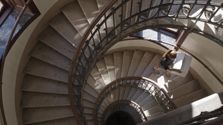 A woman carries a box up a long spiral staircase as sunlight shines in through the windows