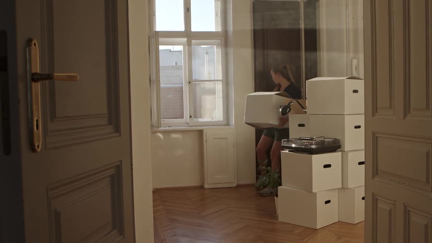A woman enters a room while carrying a cardboard box and shuts the door with her foot