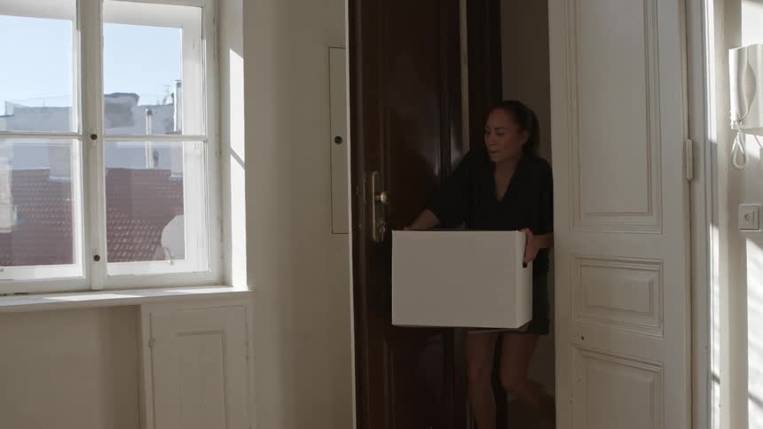 A woman opens a door to an apartment and enters carrying a white box in her hands