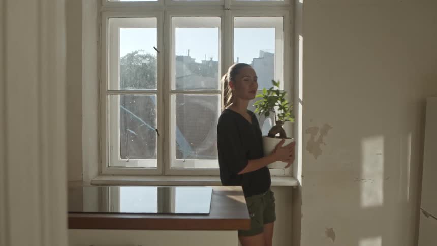 A woman picks up a plant on a window sill and carries it through the room