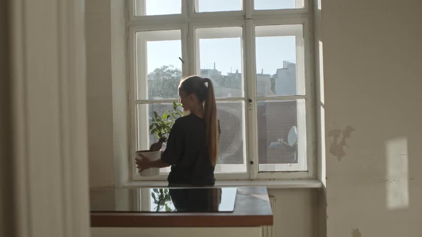 A woman places a plant on a window sill and looks outside