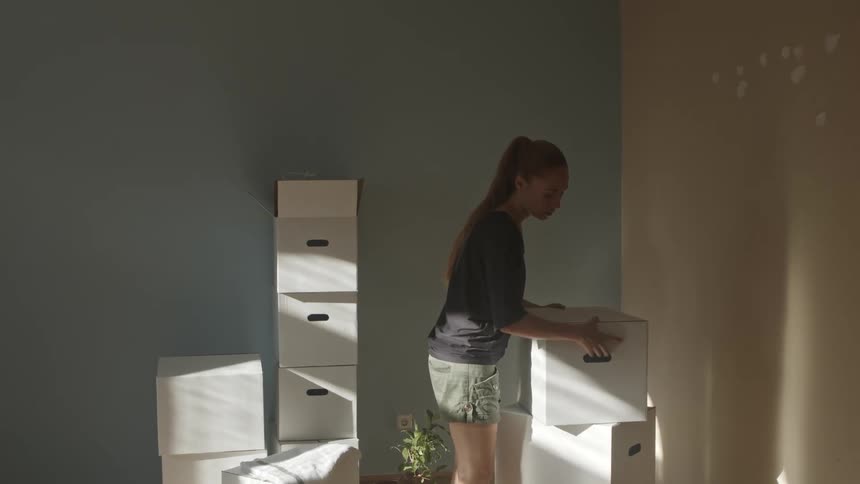 A woman approaches and lifts a cardboard box before carrying it through a house as sunlight shines in through a window