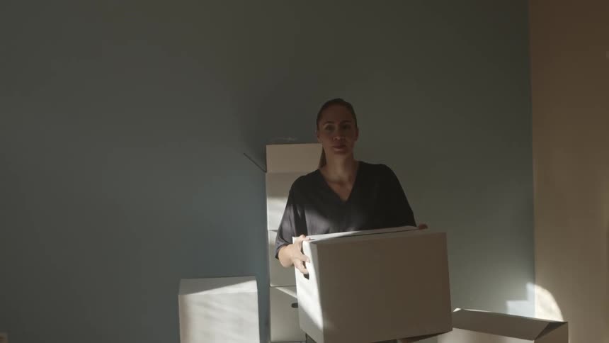 A woman lifts a large cardboard box and walks out of the room with it