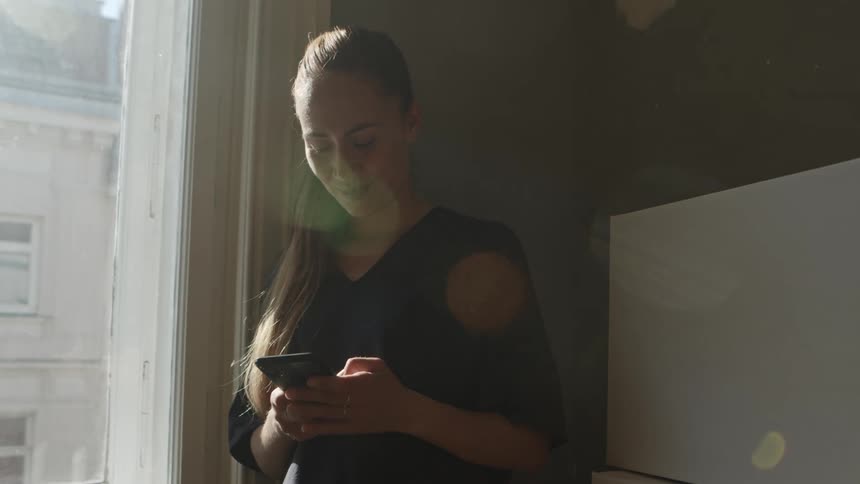 Woman with a black shirt and a brown ponytail stands by the windows and uses a smartphone in the room