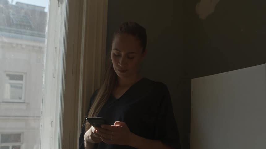 A woman stands by a window at home and looks at her cell phone