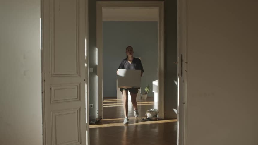 A woman carries a cardboard box through a house corridor during the day