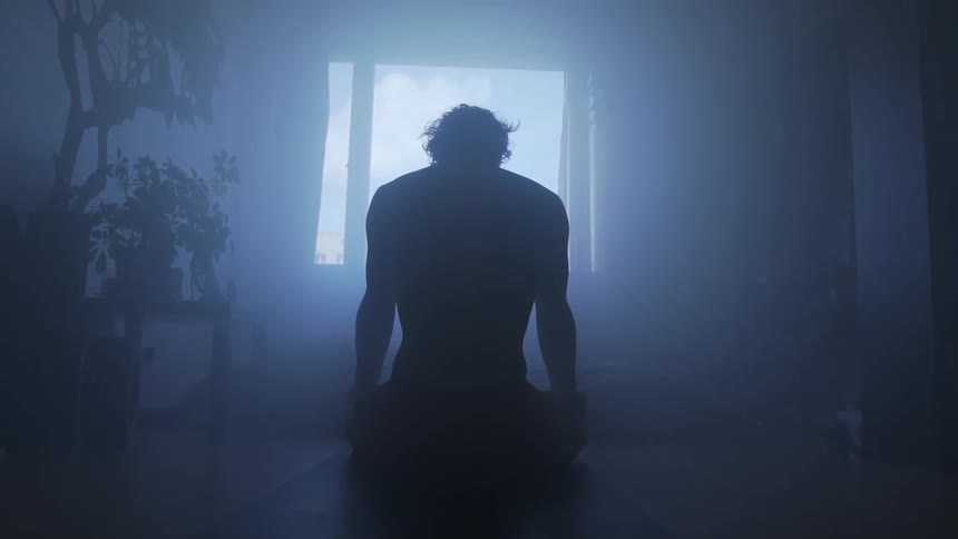 A man mediates on the floor of a dark room as sunlight shines in through the window
