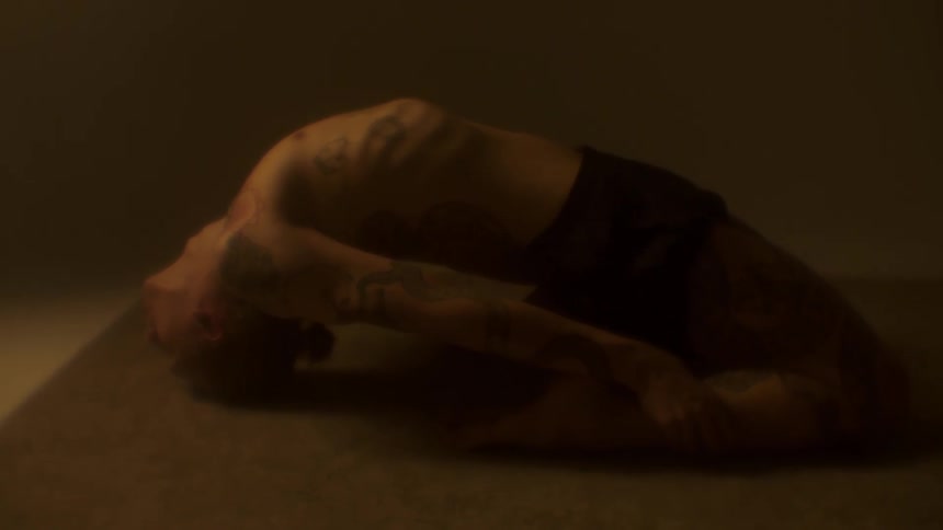 A man shirtless tattooed man holds a yoga pose on a yoga mat in an enclosed space