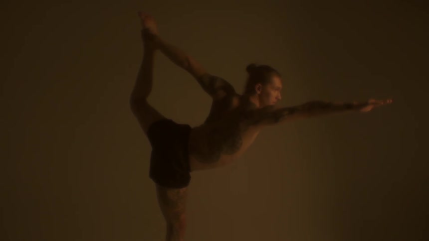 A man stretches in a yoga like pose inside of a dim lit room