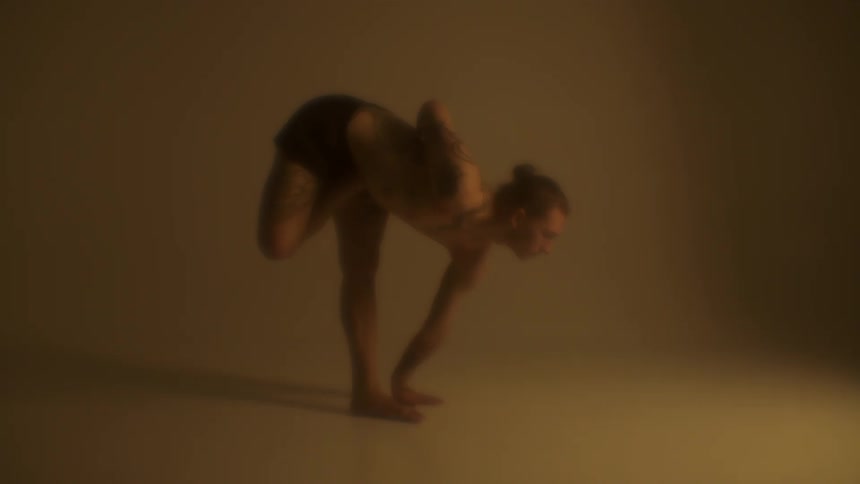 A shirtless tattooed man holds a difficult yoga position as the camera draws near him