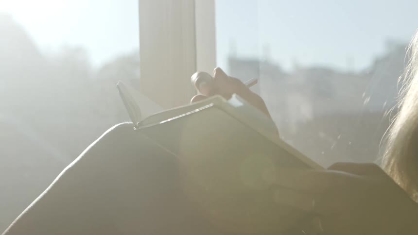 Woman with brown hair sits on windowsill and writes in a notebook