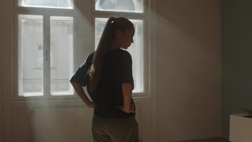 A woman puts her hands to her hips and turns in an empty room to look at moving boxes next to windows with a lot of sunlight streaming inside the apartment
