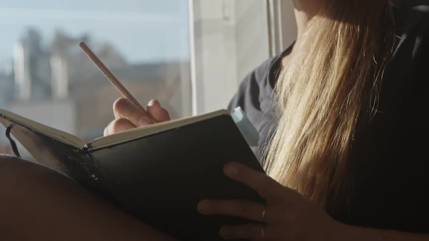 Woman with brown hair sits on windowsill and writes in a notebook
