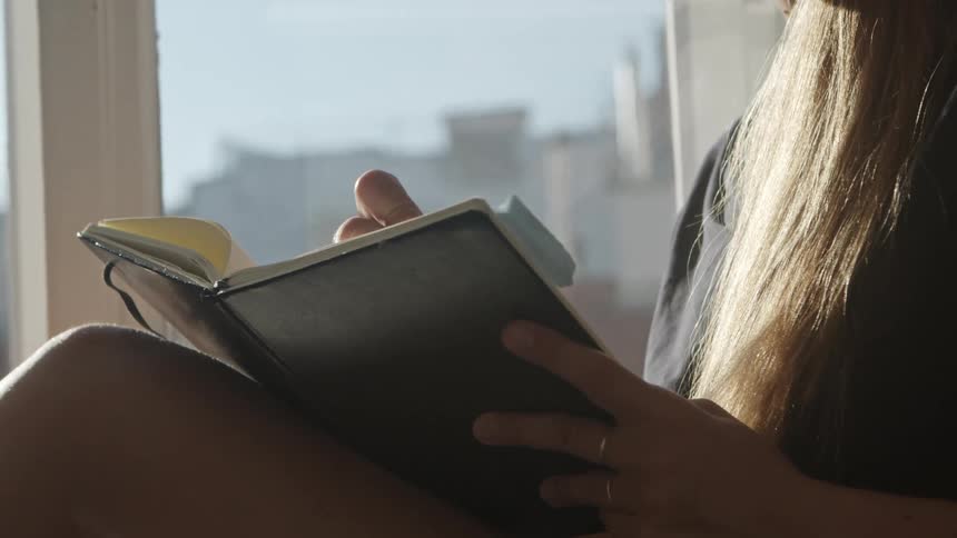 Woman with brown hair sits on windowsill and writes in a notebook