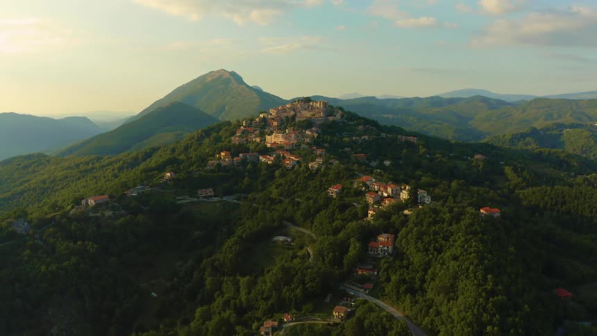 A small town is located at the top of a green hill covered in trees and shrubbery viewed from the sky