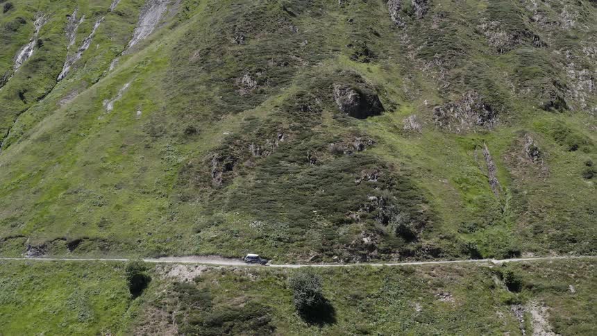 A vehicle travels along a dirt road on the side of a mountain covered in short vegetation during the day