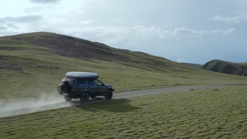 An suv drives along a remote road on hills with a landscape in the background with sun and clouds