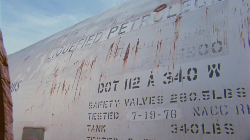 Empty old rusty petroleum oil barrels sit in front of a building surrounded by grass in daylight