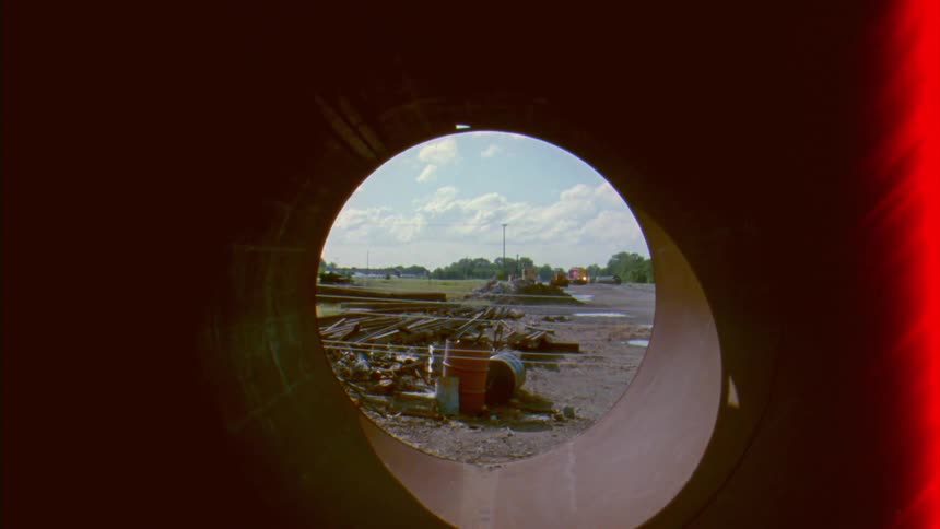An unknown person films inside of a hollow dark tube and then exits to a cloudy day surrounded by a rural area