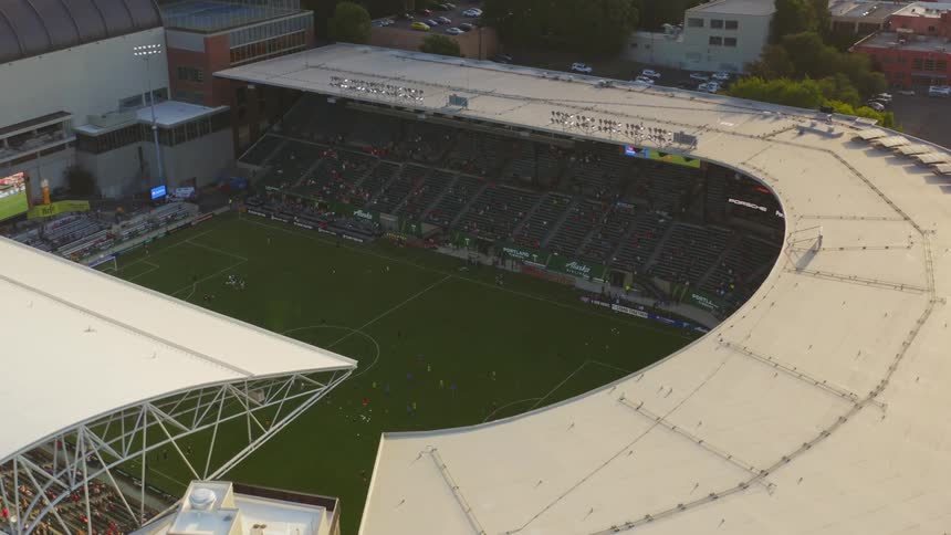 A drone flies over a stadium with a soccer field while players play and people watch on in the stands