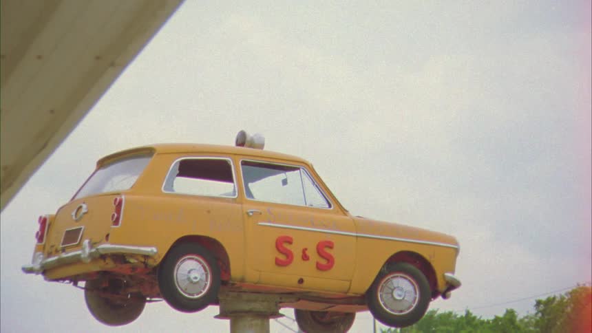 A small yellow vintage car spins on a pole outside a business district