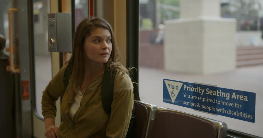 A young woman sitting in a bus traveling through a city gazes out the window before taking her smartphone from her pocket