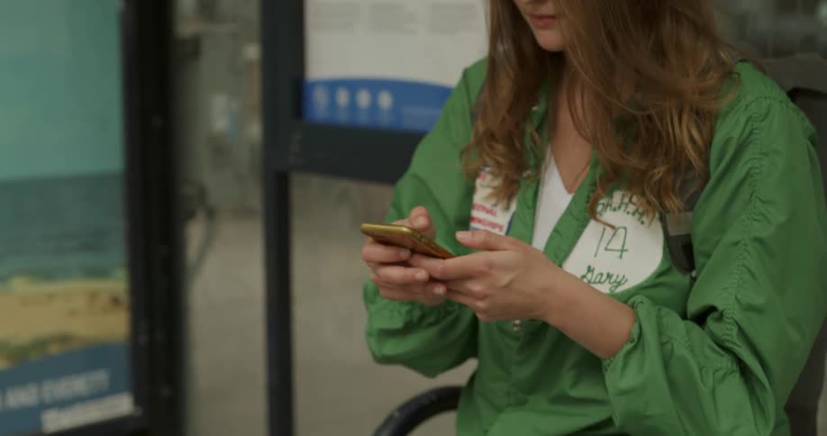A woman wearing sunglasses and a green shirt sits on a bench texting on her phone