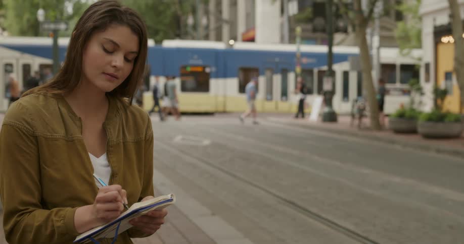 A woman writes in notebook while standing outdoors in a metropolis during the day