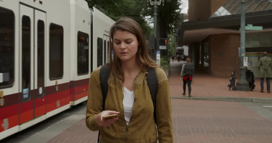 A young woman stands on a brick sidewalk in a city texting on her phone as a tram passes her on the street