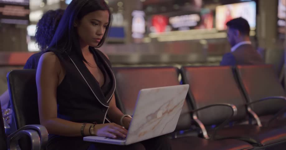 A woman sits inside of a building typing on a laptop where people are sitting in the background