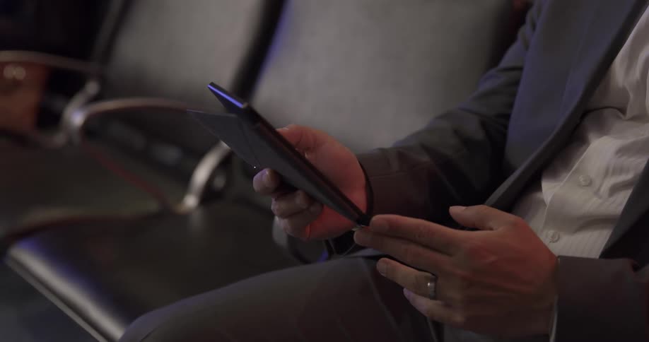 Man with a blazer and a shirt sits in a black chair and uses a smartphone in the airport terminal