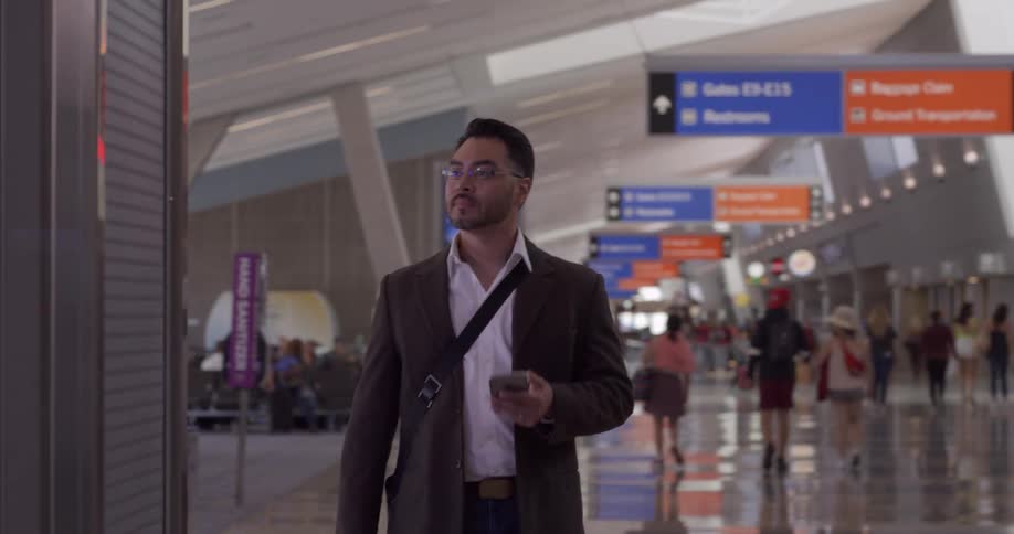 A man with glasses and a smart phone walking in a terminal with people in the background