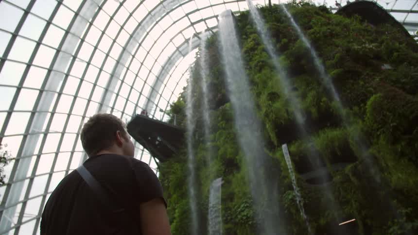 A man looks at a man made water fall in a greenhouse