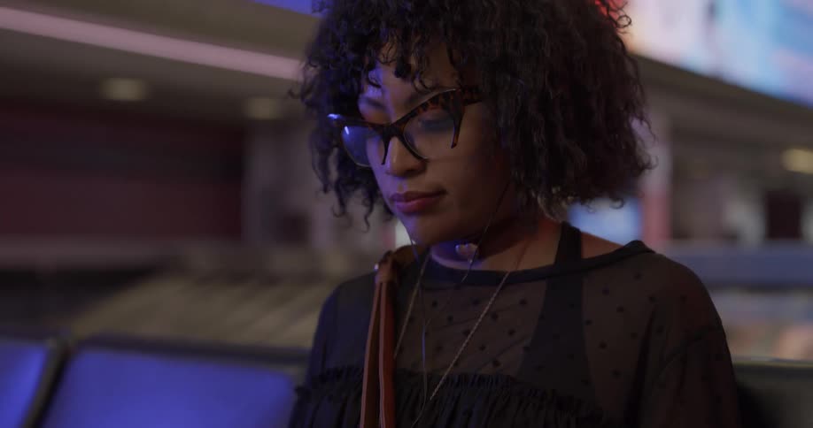 Woman with curly hair glasses and a black blouse sits in a seat and uses her smartphone in the airport terminal
