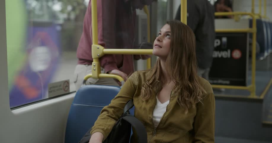 A woman sits on a train staring off outside of the window next to her smiling while a man stands behind her close to the exit on his phone