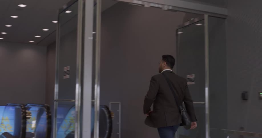A man in business casual clothing walks outdoors along a shopping center before entering a building and descending an escalator